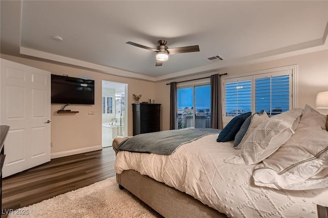 bedroom with wood finished floors, a raised ceiling, baseboards, a ceiling fan, and ensuite bath