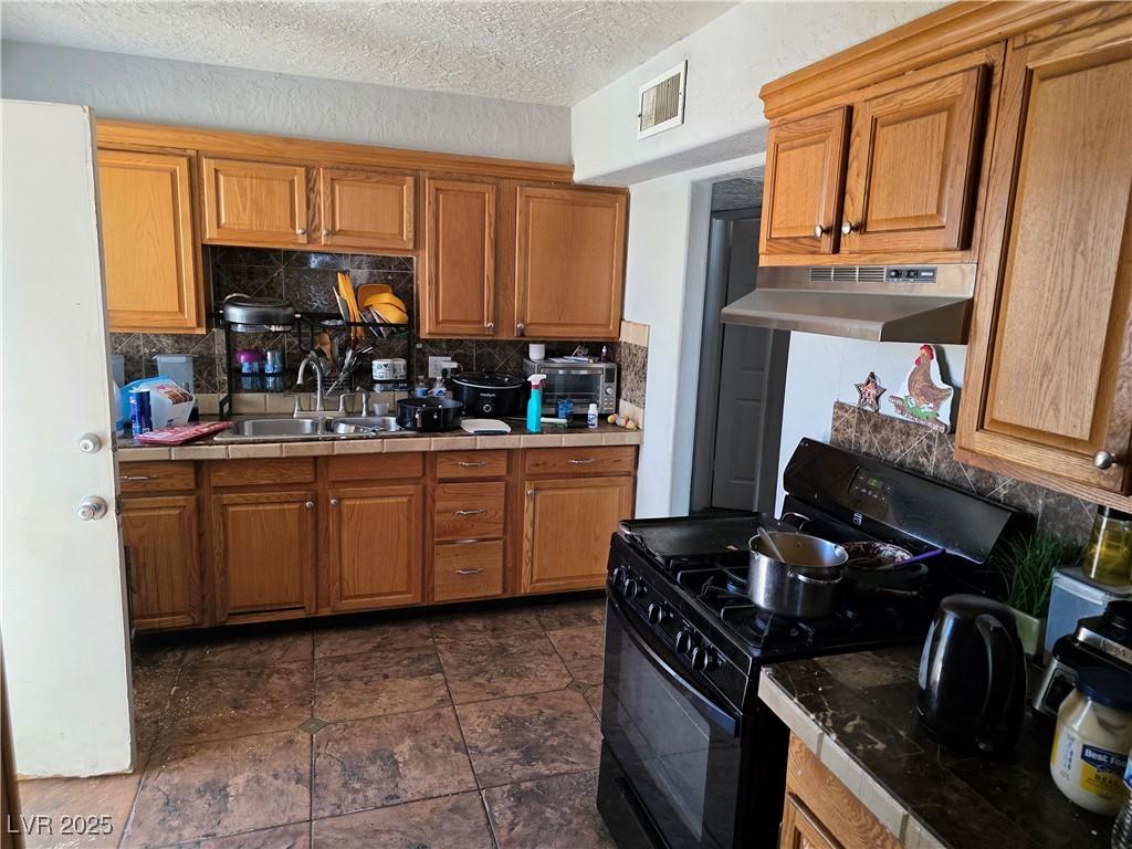 kitchen featuring gas stove, under cabinet range hood, tile countertops, a sink, and decorative backsplash