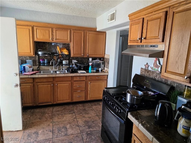 kitchen featuring gas stove, under cabinet range hood, tile countertops, a sink, and decorative backsplash