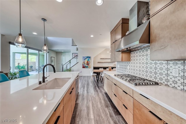 kitchen with a sink, wall chimney range hood, light countertops, light brown cabinetry, and recessed lighting