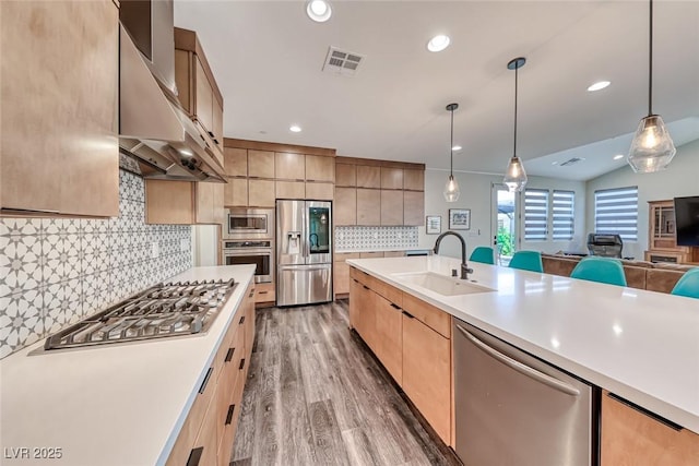 kitchen with appliances with stainless steel finishes, a sink, wall chimney exhaust hood, light countertops, and light wood-style flooring
