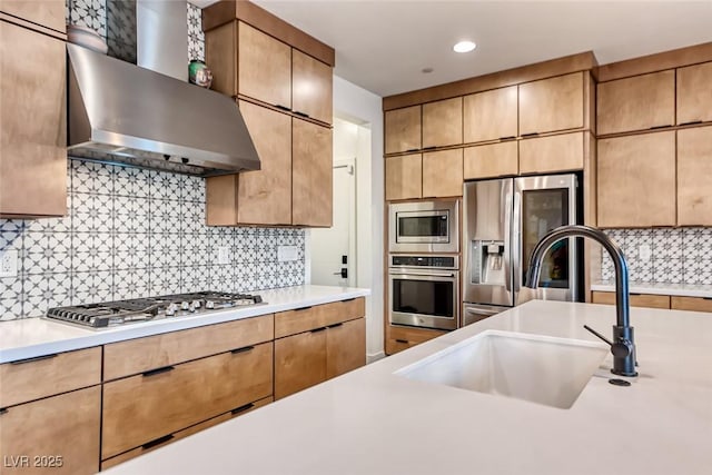 kitchen with stainless steel appliances, wall chimney range hood, a sink, light countertops, and tasteful backsplash