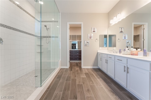 bathroom featuring a walk in shower, wood finish floors, double vanity, baseboards, and a walk in closet
