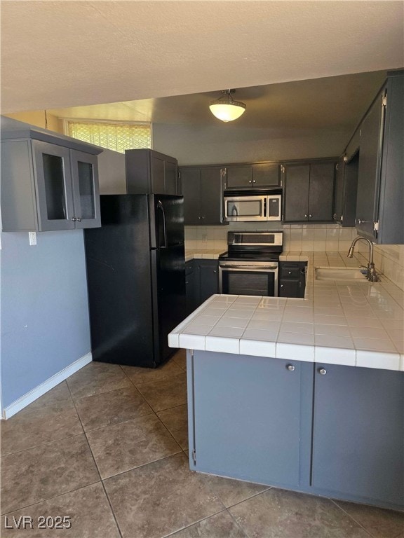 kitchen with stainless steel appliances, a sink, tile counters, a peninsula, and baseboards