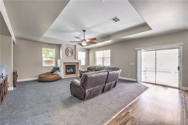 living area with a raised ceiling, baseboards, a ceiling fan, a glass covered fireplace, and wood finished floors