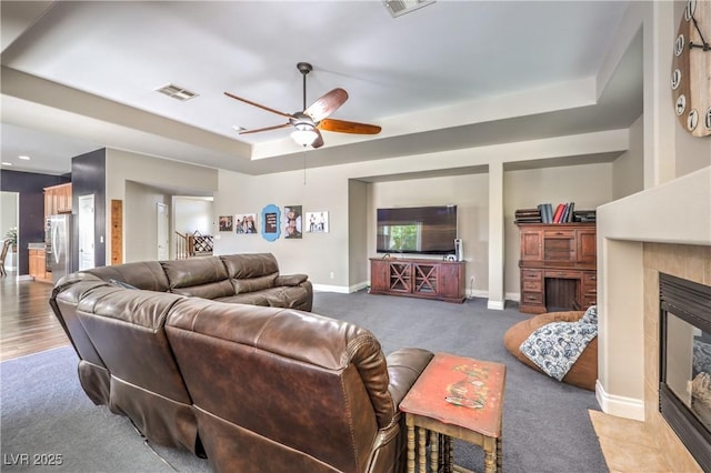 carpeted living area featuring a tray ceiling, ceiling fan, a tiled fireplace, and baseboards