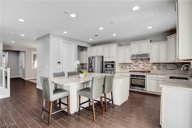 kitchen featuring appliances with stainless steel finishes, a sink, under cabinet range hood, a center island, and backsplash