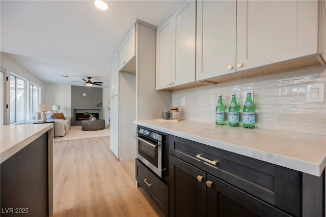 kitchen featuring oven, decorative backsplash, light countertops, a ceiling fan, and a warm lit fireplace