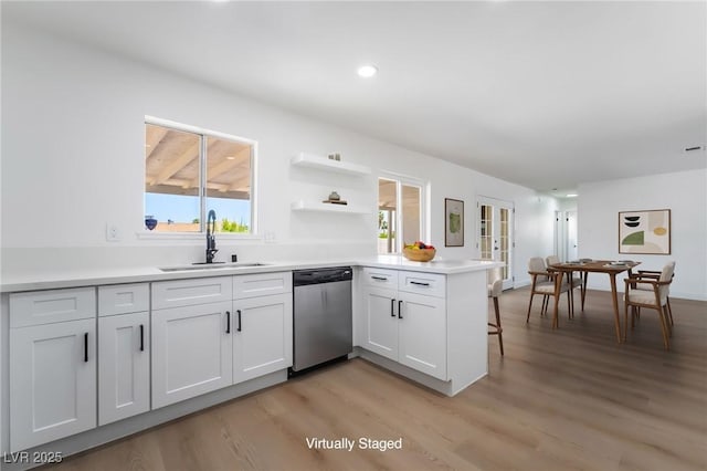 kitchen with stainless steel dishwasher, a sink, a peninsula, light wood-type flooring, and white cabinetry