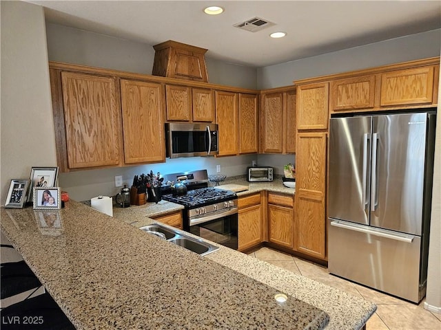 kitchen featuring appliances with stainless steel finishes, a sink, a peninsula, light tile patterned floors, and brown cabinetry