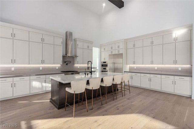 kitchen featuring wall chimney range hood, a high ceiling, stainless steel appliances, tasteful backsplash, and white cabinetry