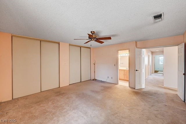 unfurnished bedroom featuring two closets, light carpet, a textured ceiling, ensuite bathroom, and a ceiling fan
