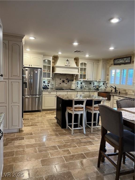 kitchen with stainless steel refrigerator with ice dispenser, custom range hood, a sink, a kitchen bar, and backsplash