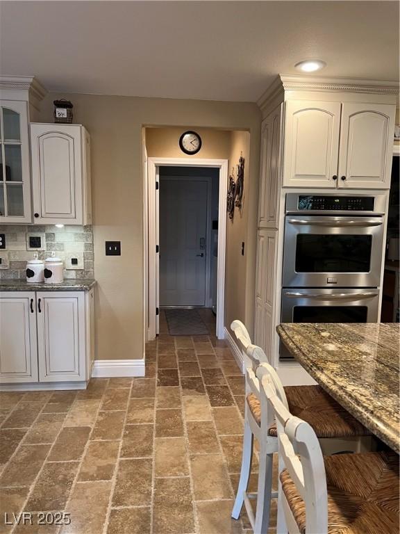kitchen featuring double oven, baseboards, dark stone countertops, glass insert cabinets, and tasteful backsplash