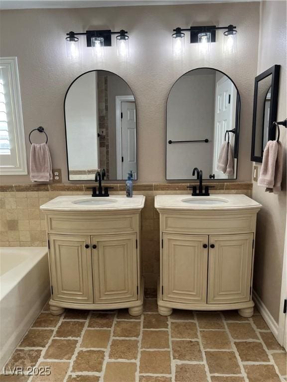bathroom featuring tile walls, two vanities, brick patterned floors, a wainscoted wall, and a tub
