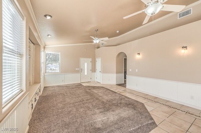 spare room featuring a ceiling fan, light tile patterned floors, crown molding, arched walkways, and a wainscoted wall