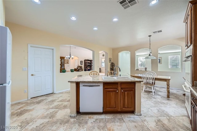 kitchen featuring a sink, arched walkways, white appliances, a center island with sink, and hanging light fixtures