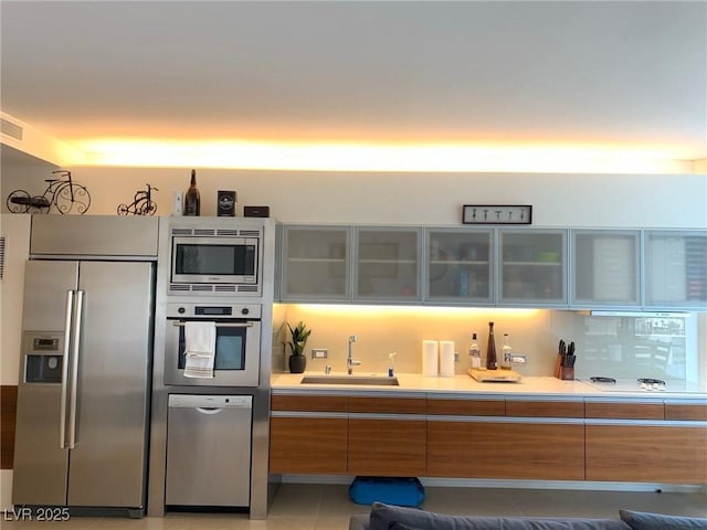 kitchen featuring stainless steel appliances, a sink, light countertops, brown cabinetry, and glass insert cabinets