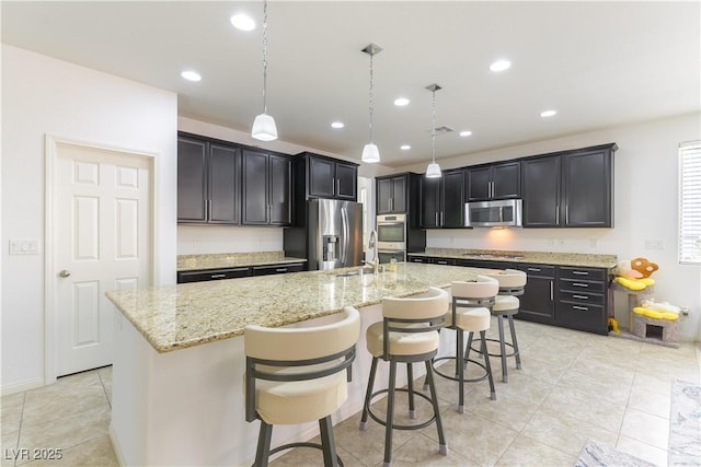 kitchen with stainless steel appliances, an island with sink, light stone countertops, light tile patterned flooring, and a breakfast bar area
