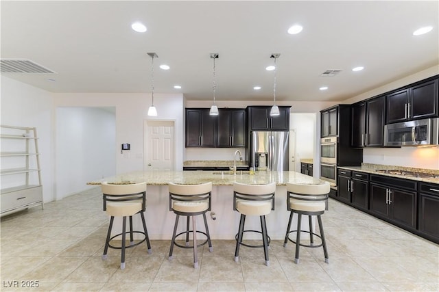 kitchen featuring appliances with stainless steel finishes, light stone counters, a kitchen bar, a center island with sink, and recessed lighting