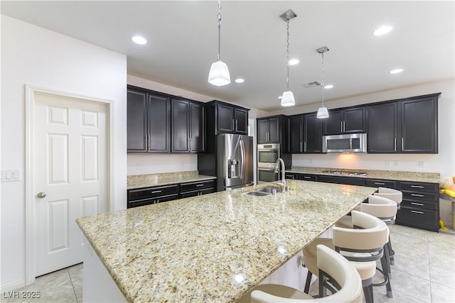 kitchen with appliances with stainless steel finishes, a sink, light tile patterned floors, light stone counters, and a center island with sink