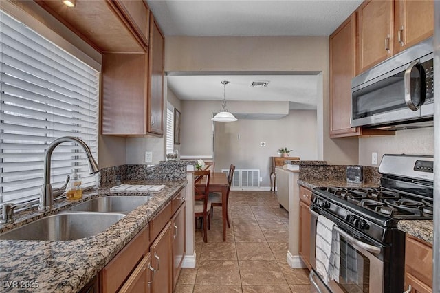 kitchen with appliances with stainless steel finishes, a sink, light tile patterned floors, dark stone counters, and brown cabinetry