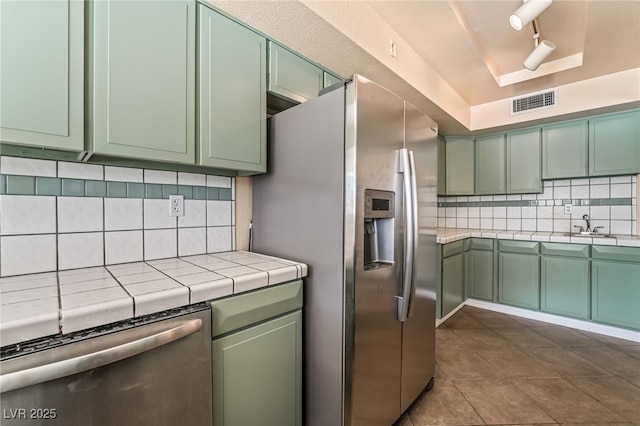 kitchen featuring green cabinetry, stainless steel appliances, tile countertops, a raised ceiling, and backsplash
