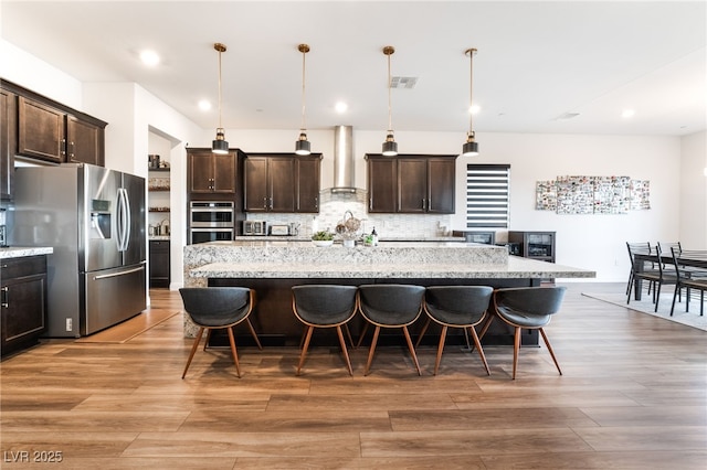 212 Harris Peak St, Las Vegas, NV 89138 - kitchen featuring stainless steel appliances, wall chimney range hood, dark brown cabinets, a kitchen bar, and light wood-style flooring