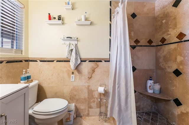 full bathroom featuring tile walls, a tile shower, toilet, vanity, and a wainscoted wall
