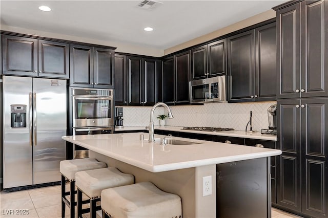 kitchen featuring stainless steel appliances, a sink, light tile patterned floors, backsplash, and a center island with sink
