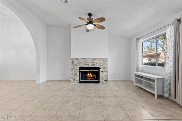 unfurnished living room featuring ceiling fan, tile patterned flooring, a stone fireplace, a textured ceiling, and arched walkways
