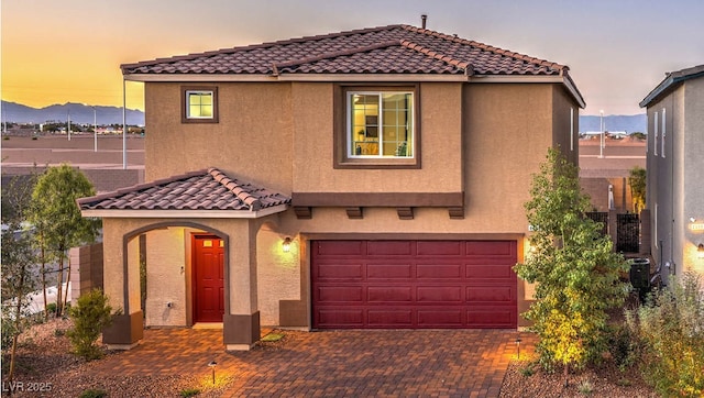 mediterranean / spanish house featuring an attached garage, stucco siding, a tiled roof, decorative driveway, and central AC
