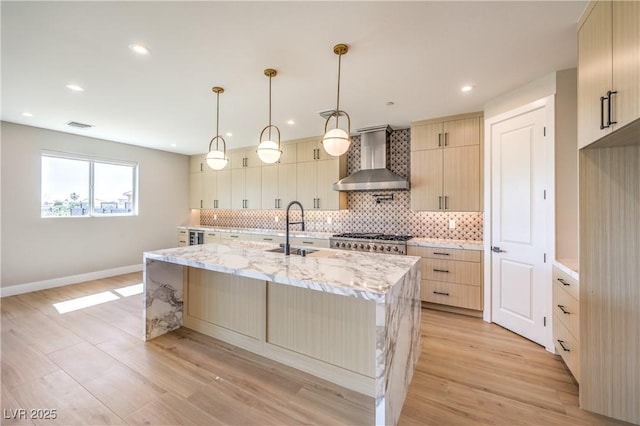 kitchen featuring wall chimney exhaust hood, a sink, backsplash, a kitchen island with sink, and light wood finished floors