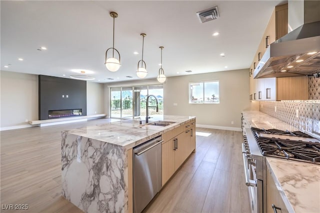 kitchen with appliances with stainless steel finishes, wall chimney range hood, a sink, light wood finished floors, and baseboards