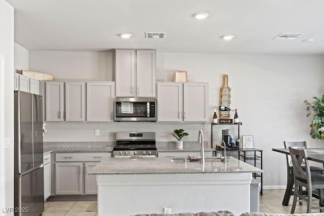 kitchen with appliances with stainless steel finishes, a sink, light tile patterned floors, light stone counters, and an island with sink