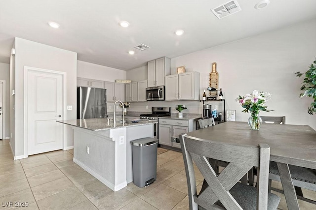 kitchen featuring appliances with stainless steel finishes, a sink, light tile patterned floors, an island with sink, and gray cabinets