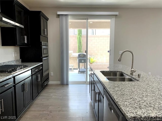 7861 Hamilton Pool Dr, Las Vegas, NV 89113 - kitchen with stainless steel appliances, a sink, light wood-type flooring, dark cabinetry, and an island with sink