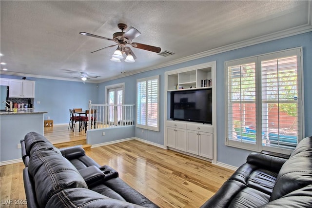 living area featuring a textured ceiling, light wood-style flooring, ceiling fan, ornamental molding, and baseboards