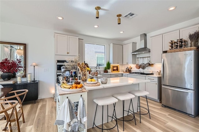 kitchen with appliances with stainless steel finishes, wall chimney range hood, a breakfast bar, light countertops, and light wood-type flooring