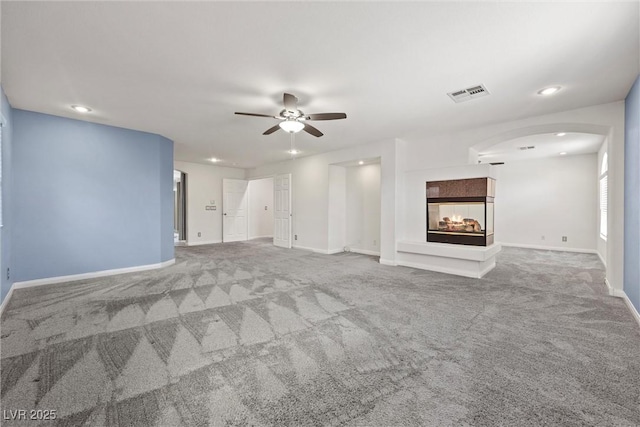unfurnished living room featuring ceiling fan, carpet, baseboards, arched walkways, and a multi sided fireplace
