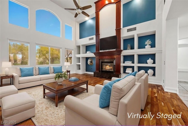 living room with a ceiling fan, built in features, a towering ceiling, wood finished floors, and a large fireplace