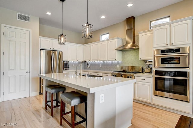 kitchen with a sink, wall chimney range hood, stainless steel appliances, decorative backsplash, and hanging light fixtures