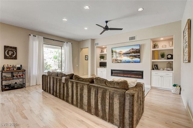 living room with light wood-style floors, a ceiling fan, a glass covered fireplace, recessed lighting, and built in shelves