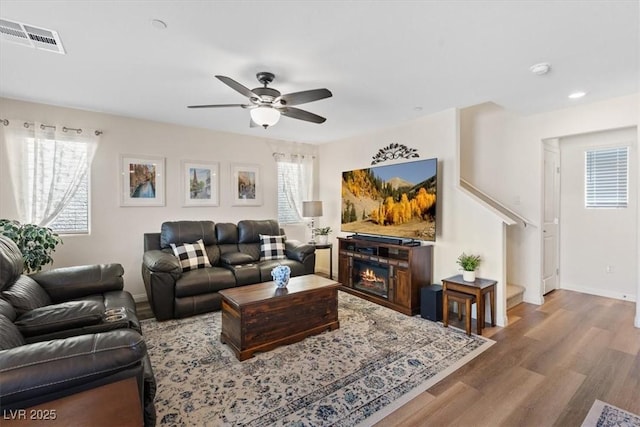 living area featuring wood finished floors, ceiling fan, stairway, baseboards, and a glass covered fireplace
