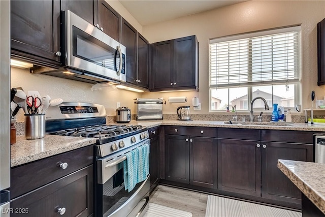 kitchen featuring stainless steel appliances, a sink, dark brown cabinets, light stone countertops, and light wood-type flooring
