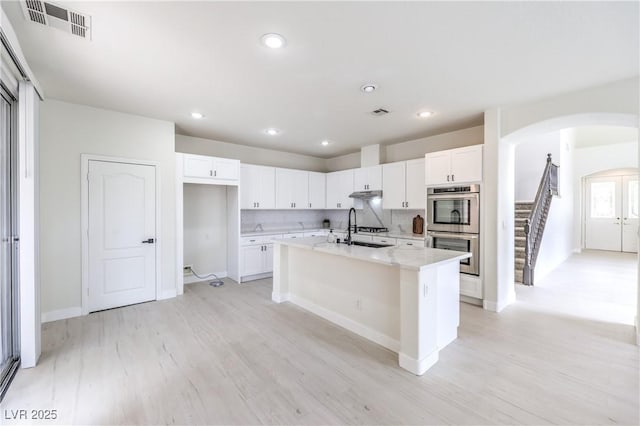 3065 Blue Monaco St, Las Vegas, NV 89117 - kitchen with double oven, arched walkways, white cabinetry, light wood-style floors, and a center island with sink
