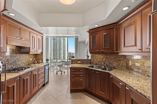 kitchen with stainless steel gas stovetop, a sink, light stone counters, decorative backsplash, and a raised ceiling