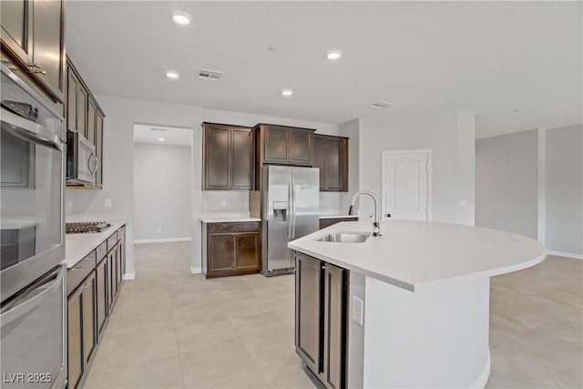 kitchen with appliances with stainless steel finishes, a sink, dark brown cabinetry, light countertops, and an island with sink