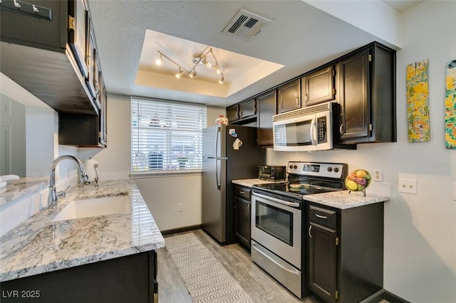 kitchen with appliances with stainless steel finishes, a sink, a tray ceiling, baseboards, and light wood-style flooring