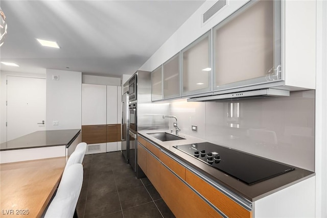 kitchen with a sink, black electric stovetop, brown cabinets, dark tile patterned floors, and glass insert cabinets